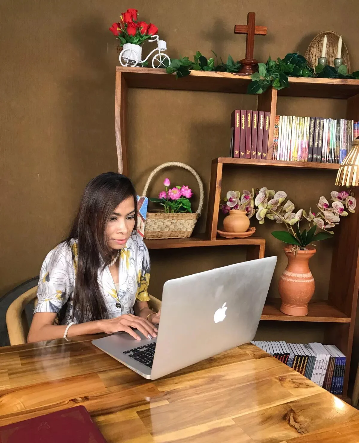 Naleng Real working at a laptop on the studio set, bookshelves and a wooden cross behind her