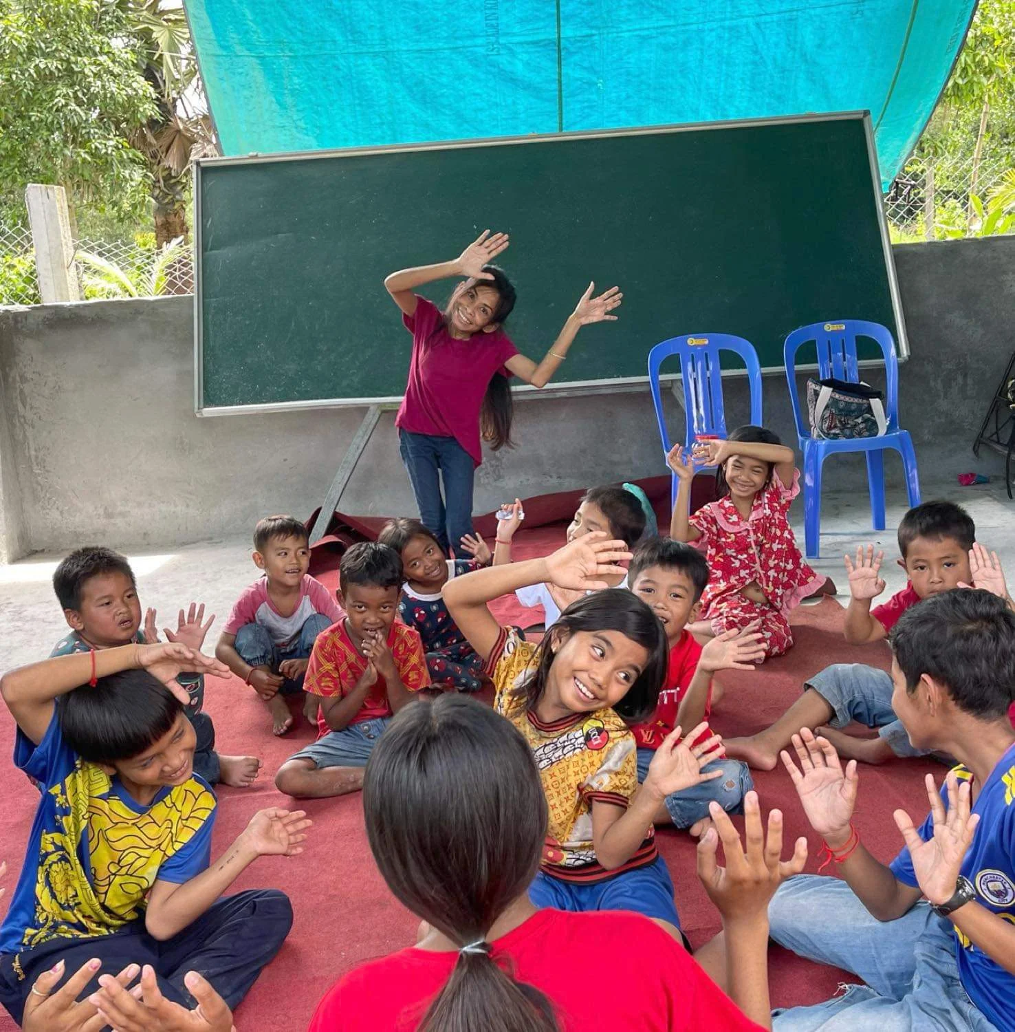 Naleng Real leading an action song for a circle of children seated on a red mat in front of a blackboard